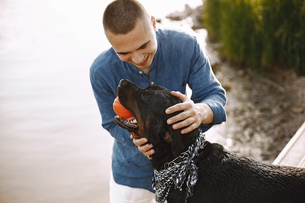 湖の近くに立っている間かわいい犬と遊ぶカジュアルな服装のハンサムな若い男性。青いシャツと白いジーンズのショートパンツを着ている男の子