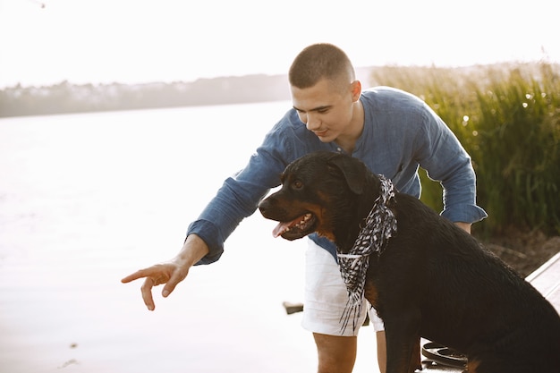 Handsome young male in casual outfit playing with cute dog while standing near the lake. Boy wearing blue shirt and white jeans shorts