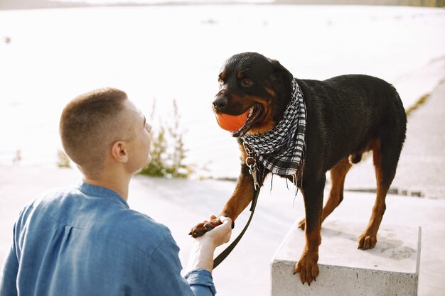 湖の近くに座ってオレンジ色のボールとかわいい犬と遊ぶカジュアルな服装のハンサムな若い男性。犬は前足を与える