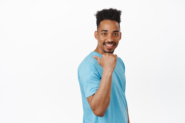 Handsome young male model model, african american guy in t-shirt pointing finger left, behind his shoulder, smiling and looking happy, invite to event on white