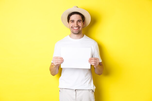 笑顔の夏の帽子のハンサムな若い男性観光客、あなたのサインのための空白の紙を持って、黄色の背景の上に立って