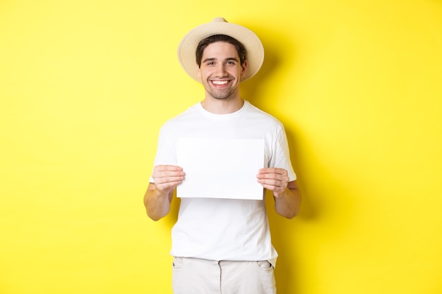여름 모자를 쓰고 웃고 있는 잘생긴 젊은 남성 관광객, 표지판을 위한 빈 종이를 들고 노란색 배경 위에 서 있습니다