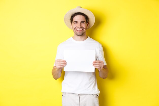 Handsome young male tourist in summer hat smiling, holding blank piece of paper for your sign, standing over yellow background