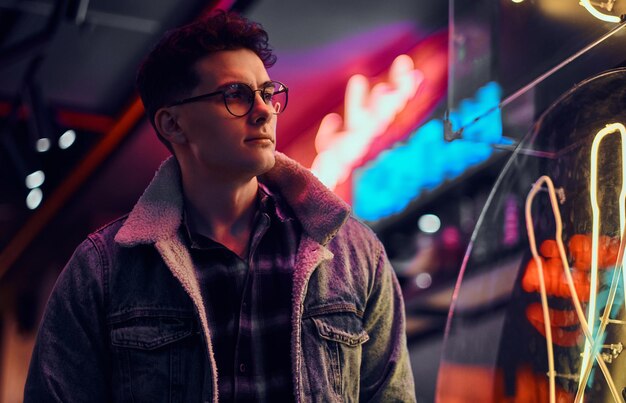 A handsome young man fashionably dressed standing in the street at night. Illuminated signboards, neon, lights.