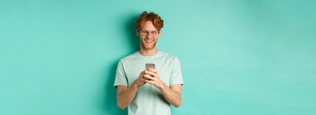 Handsome young man in glasses with red messy hair reading message on mobile phone smiling and lookin