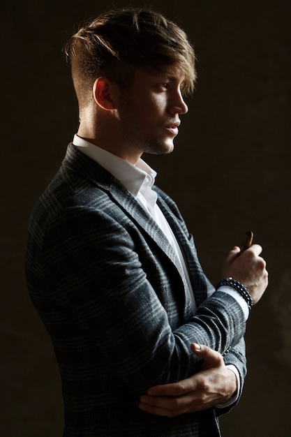 Free photo handsome young man in grey suit stands with a cigar