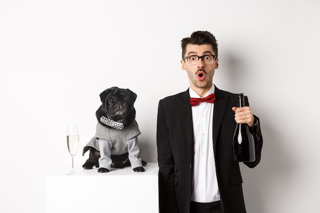 Handsome young man and his puppy celebrating New Year holiday, black pug and dog owner standing in suits, guy holding champagne, white background.