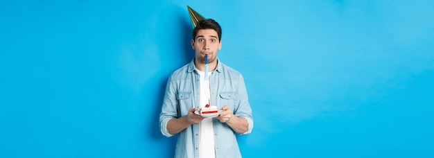 Handsome young man holding birthday cake wearing party hat and blowing whistle standing over blue ba