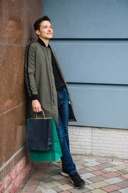 Handsome young man holding colorful shopping bags leaning on wall looking away