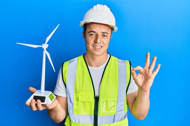 Handsome young man holding solar windmill for renewable electricity doing ok sign with fingers smiling friendly gesturing excellent symbol