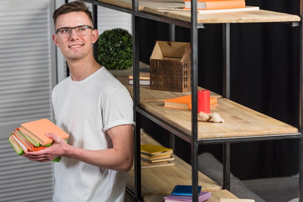 Handsome young man holding stack of books looking away standing near the shelf