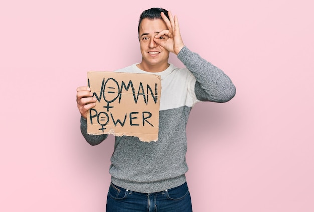 Handsome young man holding woman power banner smiling happy doing ok sign with hand on eye looking through fingers
