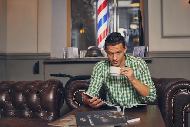 Красивый молодой человек в парикмахерской ждет мастера в зале ожидания, попивая кофе. он смотрит фотографии в модном журнале