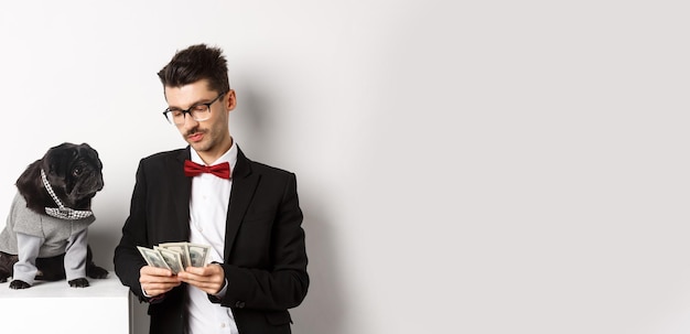 Бесплатное фото Красивый молодой человек в костюме стоит рядом с черным мопсом в костюме и считает деньги, работая на вечеринках