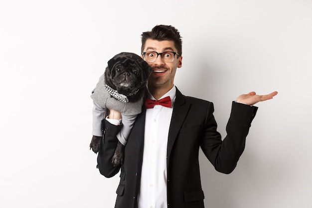 Handsome young man introducing pet product on hand, holding cute black dog on shoulder and smiling, showing something on white background