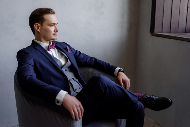 Handsome young man is sitting in the armchair in the room, dressed in the fashionable suit