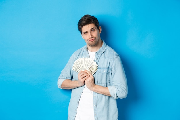 Handsome young man keeping money to himself, smiling and looking greedy, standing over blue background.