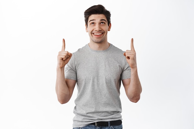 Handsome young man looking up and pointing at top copy space with dreamy face smiling pleased found interesting promo offer showing best deal product on sale white background