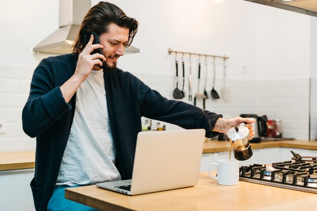 Handsome young man pouring coffee in mug talking on mobile phone with laptop on kitchen counter