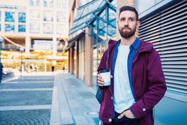 紫のジャケットを着たハンサムな若い男がコーヒーを飲んでいる 路上でコーヒーを飲んでいる男 アメリカンやラテを手にした紙カップを握っている男 ストリートスタイル