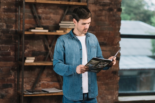 Handsome young man reading magazine standing in front of book shelf