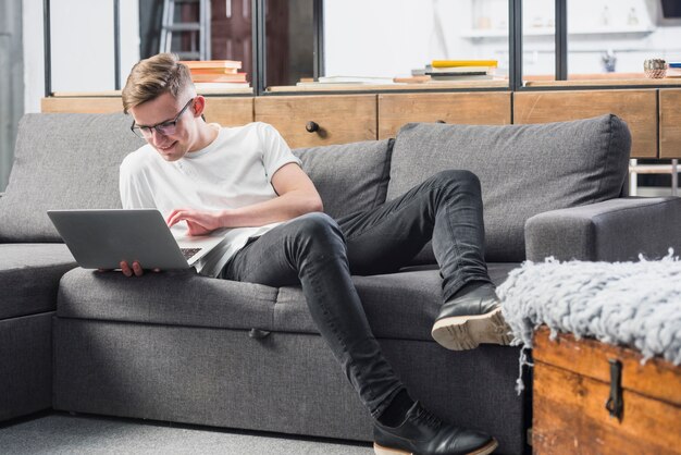 Красивый молодой человек, расслабляющий на диване, используя ноутбук дома