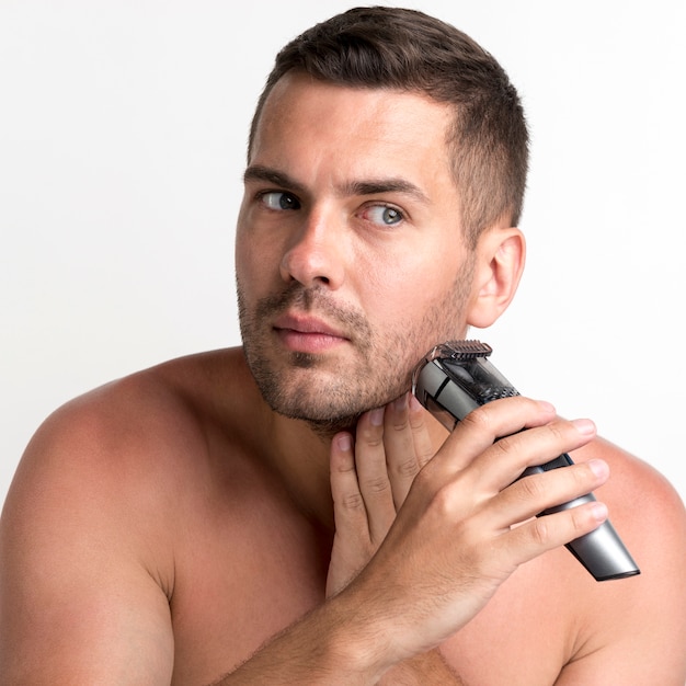 Handsome young man shaving with trimmer on white backdrop