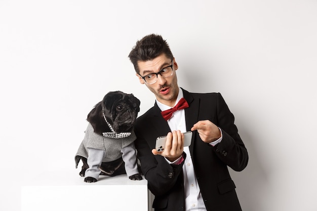 Handsome young man showing something on mobile phone to his dog. Owner shopping online with pet, standing in costumes over white background