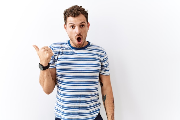 Handsome young man standing over isolated background surprised pointing with hand finger to the side open mouth amazed expression