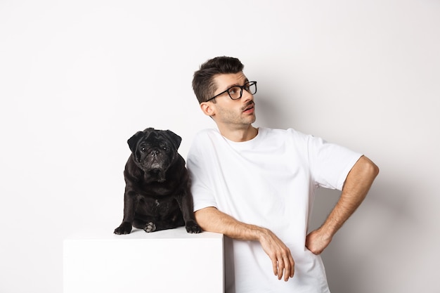 Handsome young man standing near cute black pug, looking right with arrogant expression, standing over white background