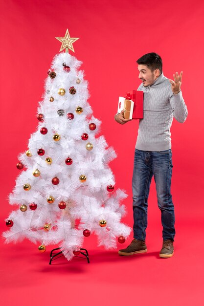 Handsome young man standing near the decorated white New Year tree and holding his gifts