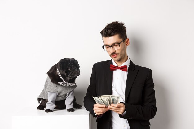 Handsome young man in suit standing near black pug in costume and counting money, working on parties, posing over white background.