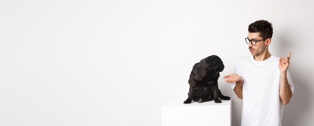 Foto gratuita giovane bello che insegna i comandi del cane che parlano con il simpatico carlino nero in piedi su sfondo bianco