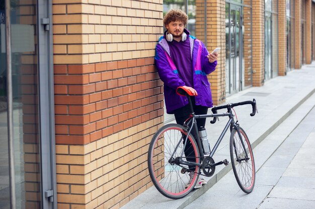 彼の横にある彼の自転車の近くに立っている間携帯電話とヘッドフォンを使用してハンサムな若い男