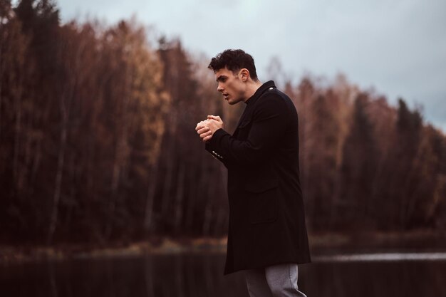 Красивый молодой человек в черном пальто греет руки у озера в осеннем лесу.