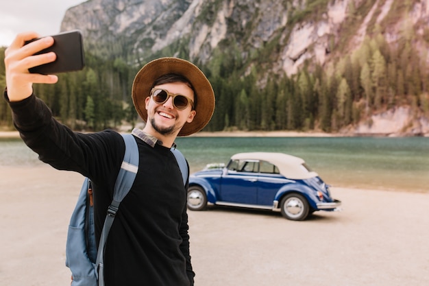 Handsome young man wearing brown hat and sunglasses taking picture of himself standing in front of mountain lake in italy