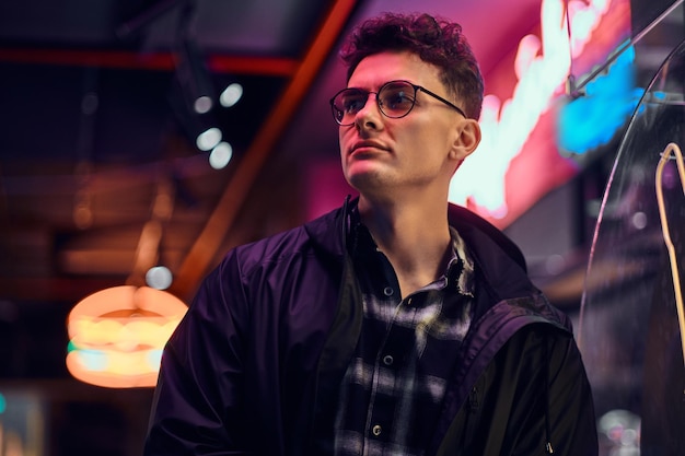 A handsome young man wearing sports hoodie standing in the street at night. Illuminated signboards, neon, lights.