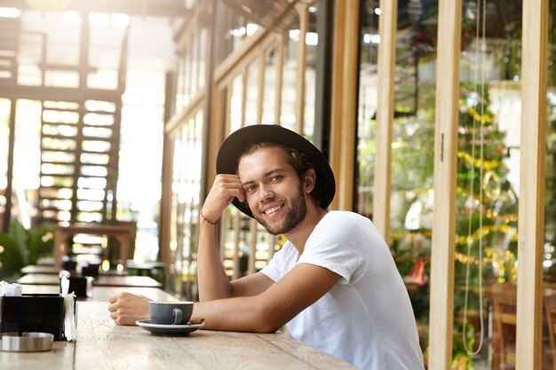 コーヒーショップの木製のテーブルに座っているトレンディな帽子をかぶっているハンサムな若い男