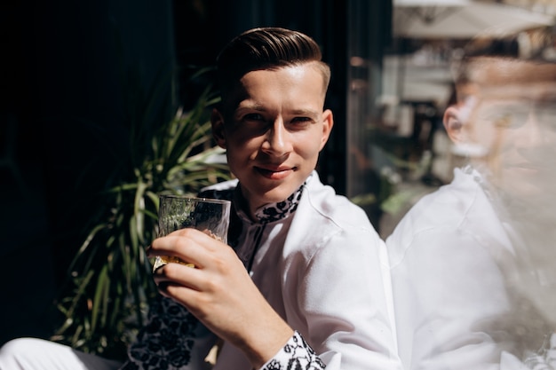 Il giovane bello in vestito bianco con ricamo si siede su un davanzale prima della finestra luminosa