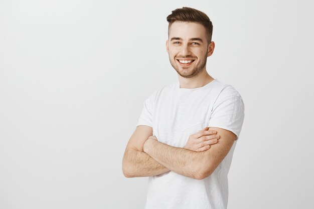 Handsome young man in white t-shirt, cross arms chest and smiling pleased