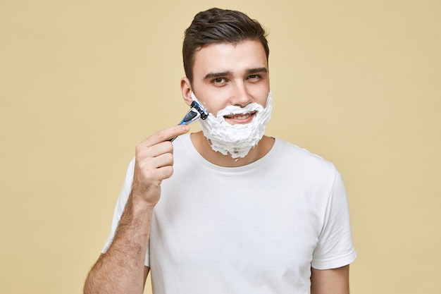 Красивый молодой человек в белой футболке, держащий бритву, бреет бороду против волокон, чтобы избежать раздражения кожи с улыбкой, заботясь о своей внешности. Мужественность, стиль и красота