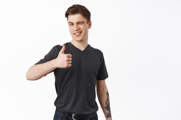 Handsome young man winks and shows thumbs up, approve and likes, recommend and leave positive feedback, praise choice, guarantee quality, white background