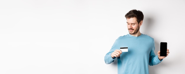 Free photo handsome young man with beard showing empty smartphone screen and looking at plastic credit card sta