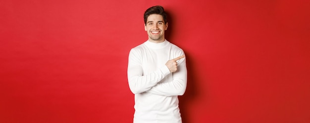 剛毛のハンサムな若い男、白いセーターを着て、右上隅に指を指して、笑顔、クリスマスバナーを表示、赤い背景の上に立って Premium写真