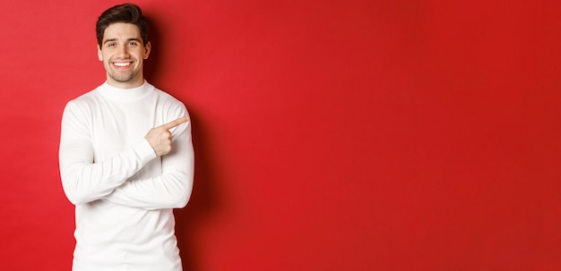 Free photo handsome young man with bristle, wearing white sweater, pointing fingers at upper right corner and smiling, showing christmas banner, standing over red background.