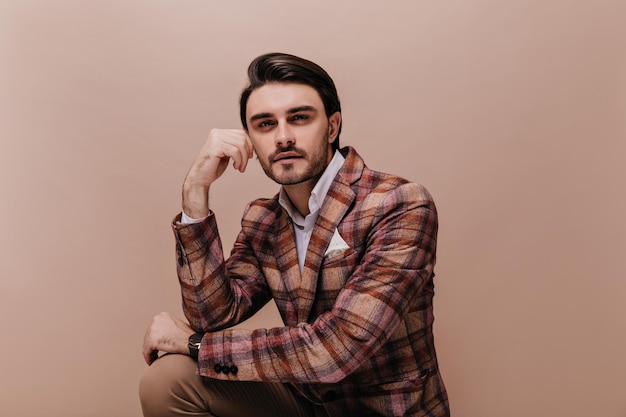 Free photo handsome young man with brunette hair and bristle sitting against beige pastel background and looking straight forward model wearing retro plaid jacket