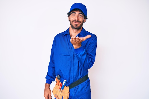 Free photo handsome young man with curly hair and bear weaing handyman uniform looking at the camera blowing a kiss with hand on air being lovely and sexy love expression