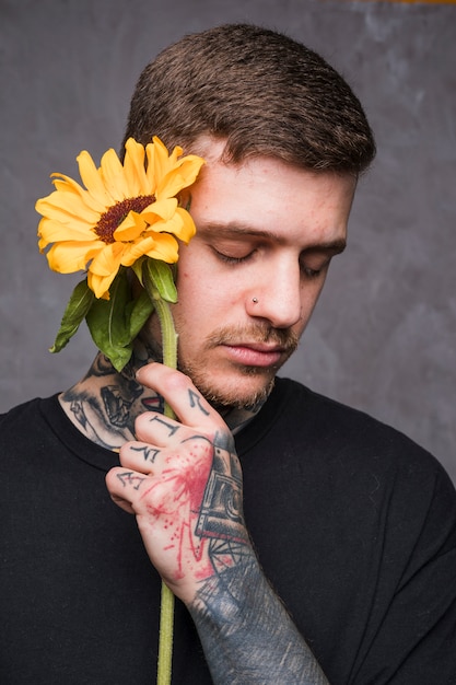 Free photo handsome young man with nose pierced holding sunflower in hand against grey backdrop