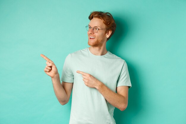 赤い髪とあごひげを持つハンサムな若い男、眼鏡とTシャツを着て、面白がって左を指して見て、コピースペース、ミントの背景の広告をチェックします。