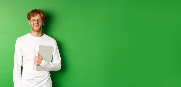 Free photo handsome young man with red hair wearing glasses and longsleeve tshirt holding laptop on green backg